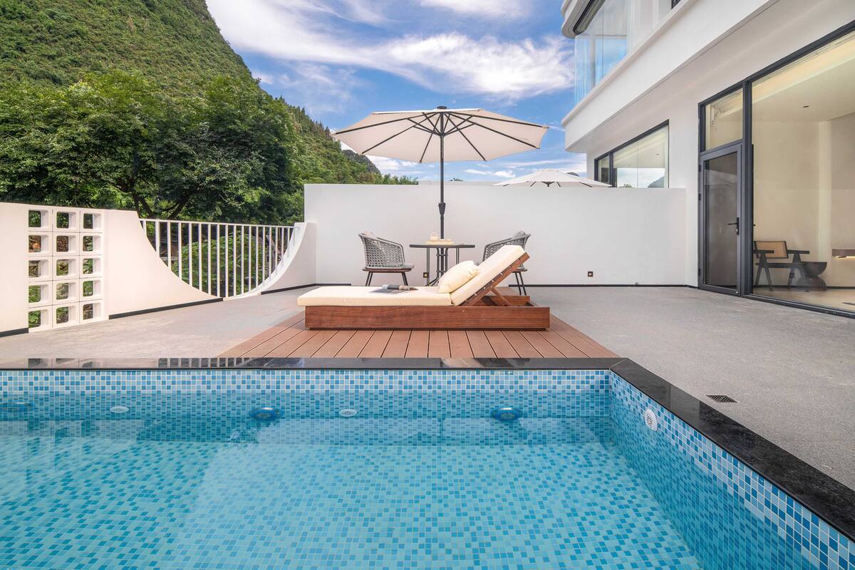 A modern outdoor pool area with a lounge chair and two chairs under a large umbrella, surrounded by a white wall and a wooden deck.
