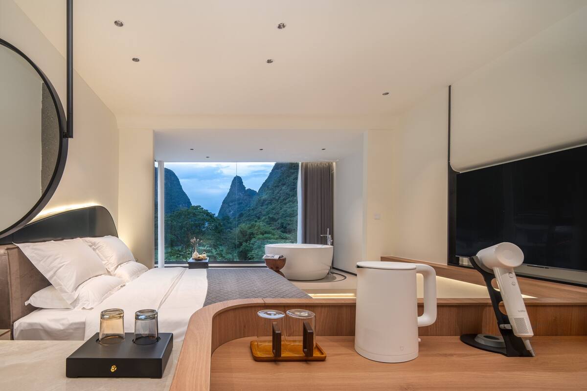A modern hotel room with a large bed, a bathtub, a desk with a coffee maker, and a view of mountains.