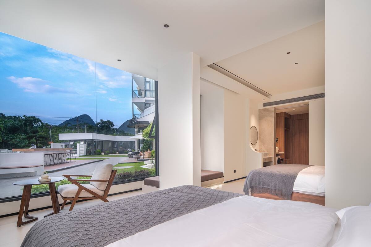 A modern hotel room with a large window offering a view of a landscaped garden and a modern building. The room includes a bed, a seating area with a table, and a walk-in closet.