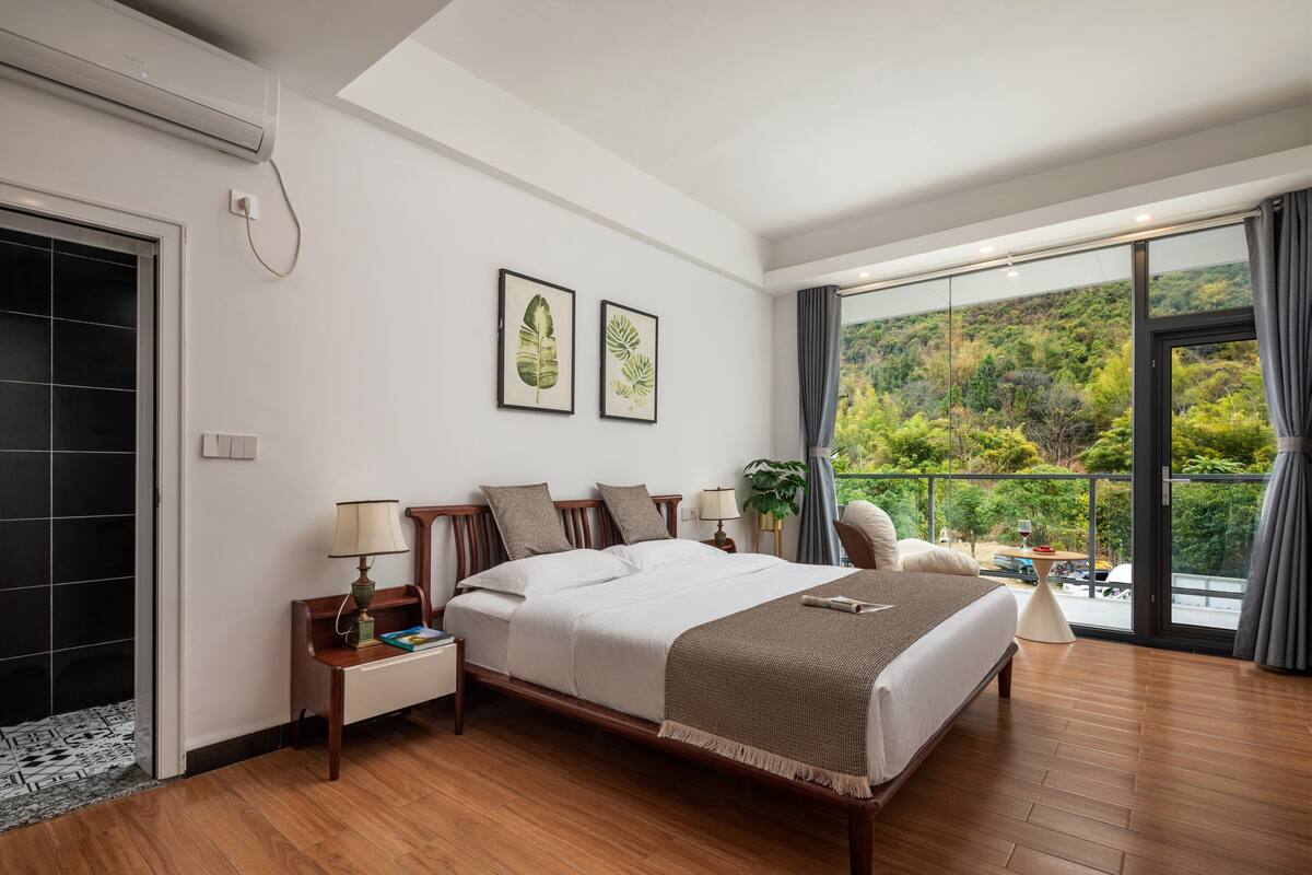 A modern hotel room with a large bed, wooden flooring, a balcony with a view of greenery, and a large window with curtains.