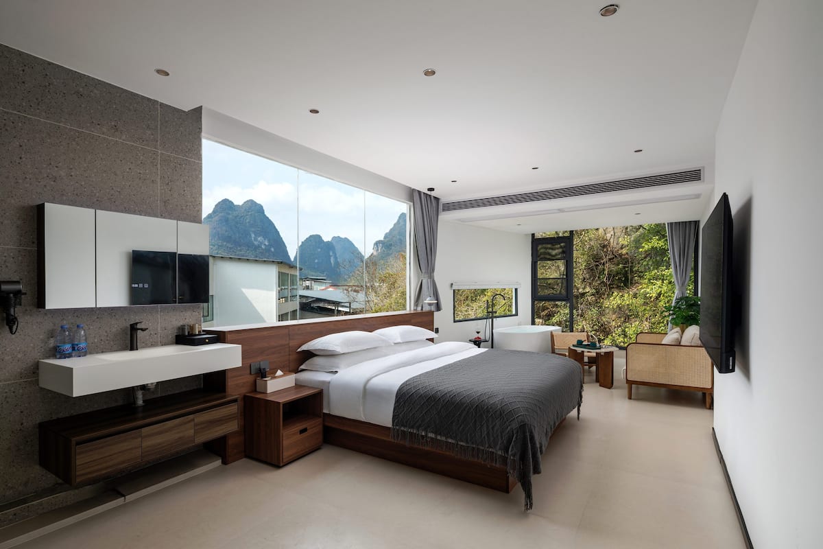 A modern bedroom with a large bed, a TV, a vanity, and a view of mountains.