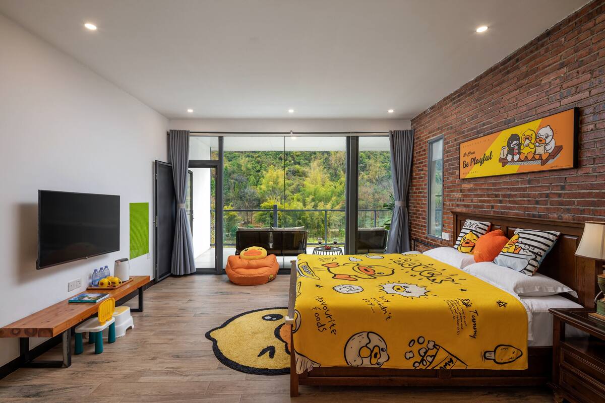 A bedroom with a large bed, a TV, a bench, and a view of greenery outside.