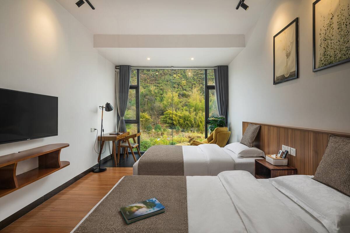 A modern hotel room with two beds, a flat-screen TV, a desk, and a view of a forest through large windows.