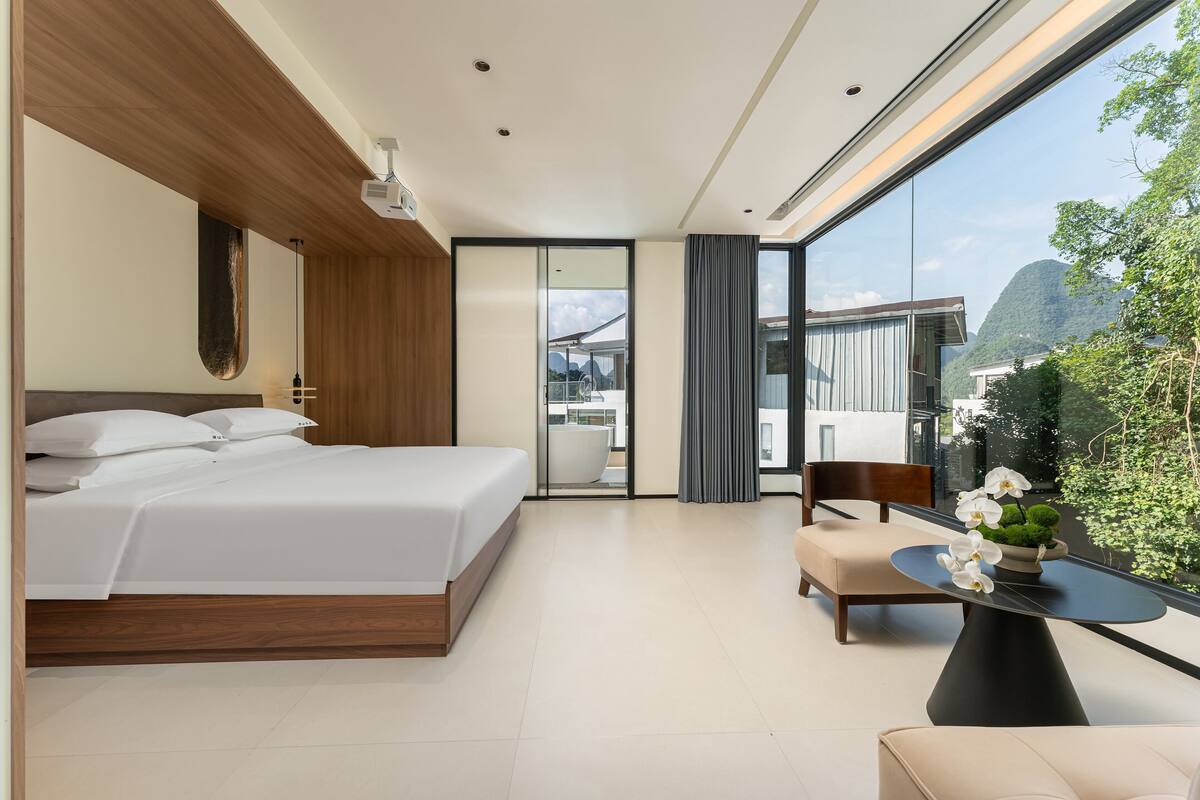 A modern hotel room with a large bed, a seating area with a table, and a view of a mountain and greenery.