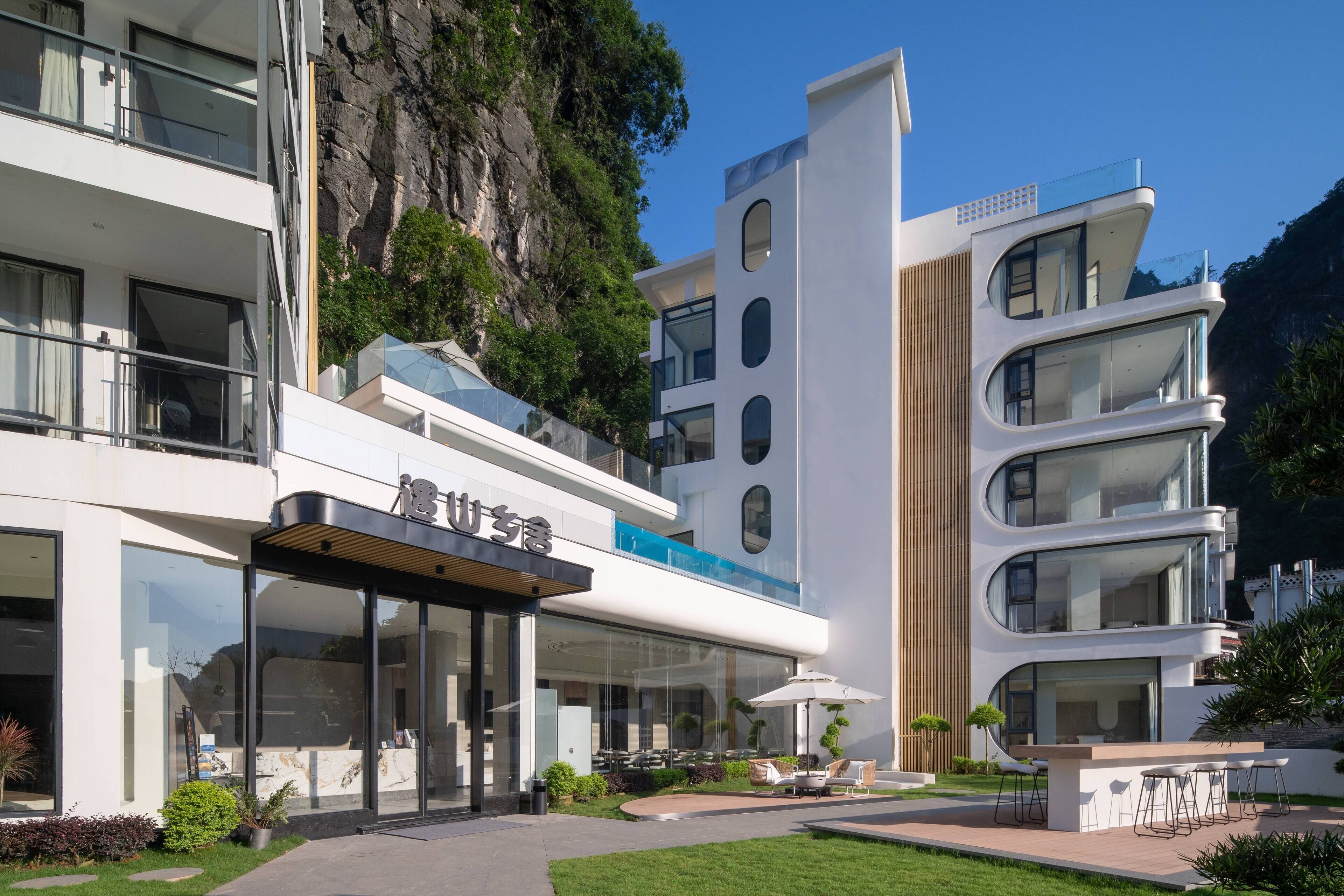 A modern hotel with a glass facade, outdoor seating area, and a view of a rocky cliff.