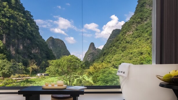 A room with a view of a mountainous landscape, a table with a tea set, and a white chair with a towel. - Karst Scenic Hotel (Guilin)