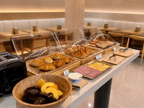 Um buffet de café da manhã com diversos pães, frios e uma salada de frutas.
