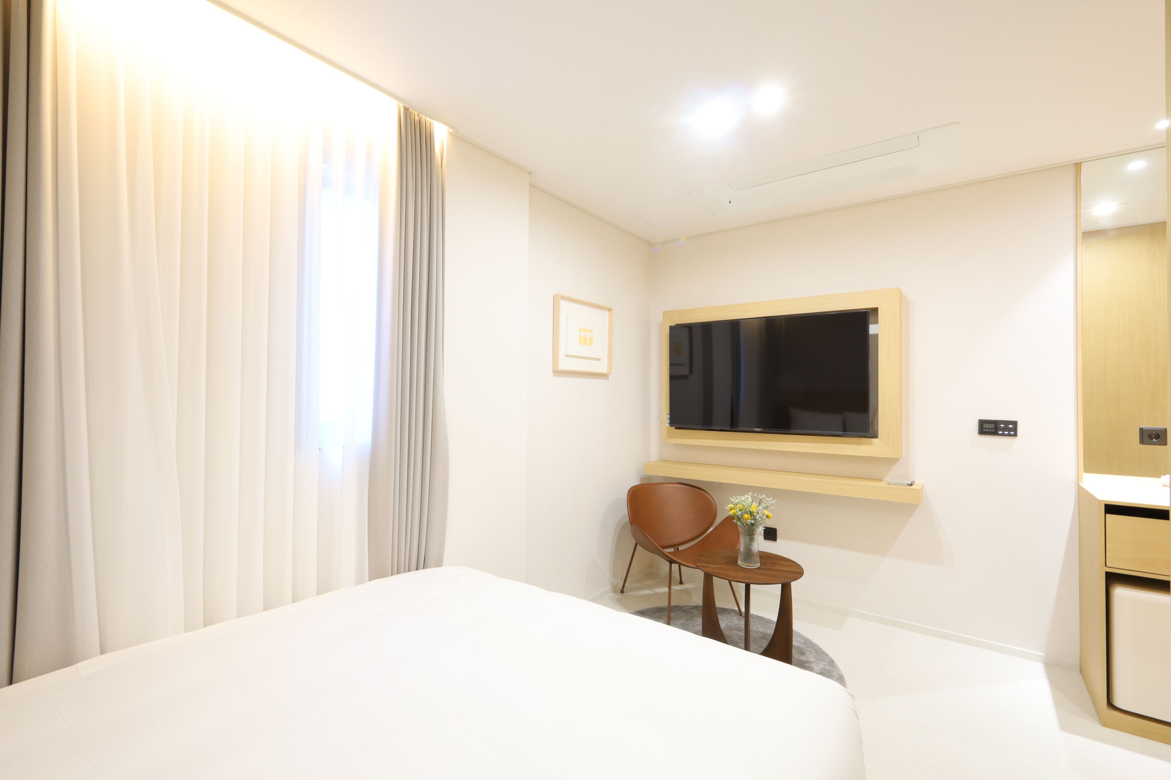 Una habitación de hotel con una cama, un televisor montado en la pared, una mesita con un florero y una silla.