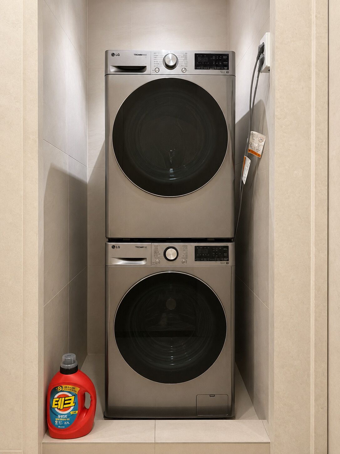 A compact laundry room with two stacked front-loading washing machines, a power outlet, and a bottle of detergent.