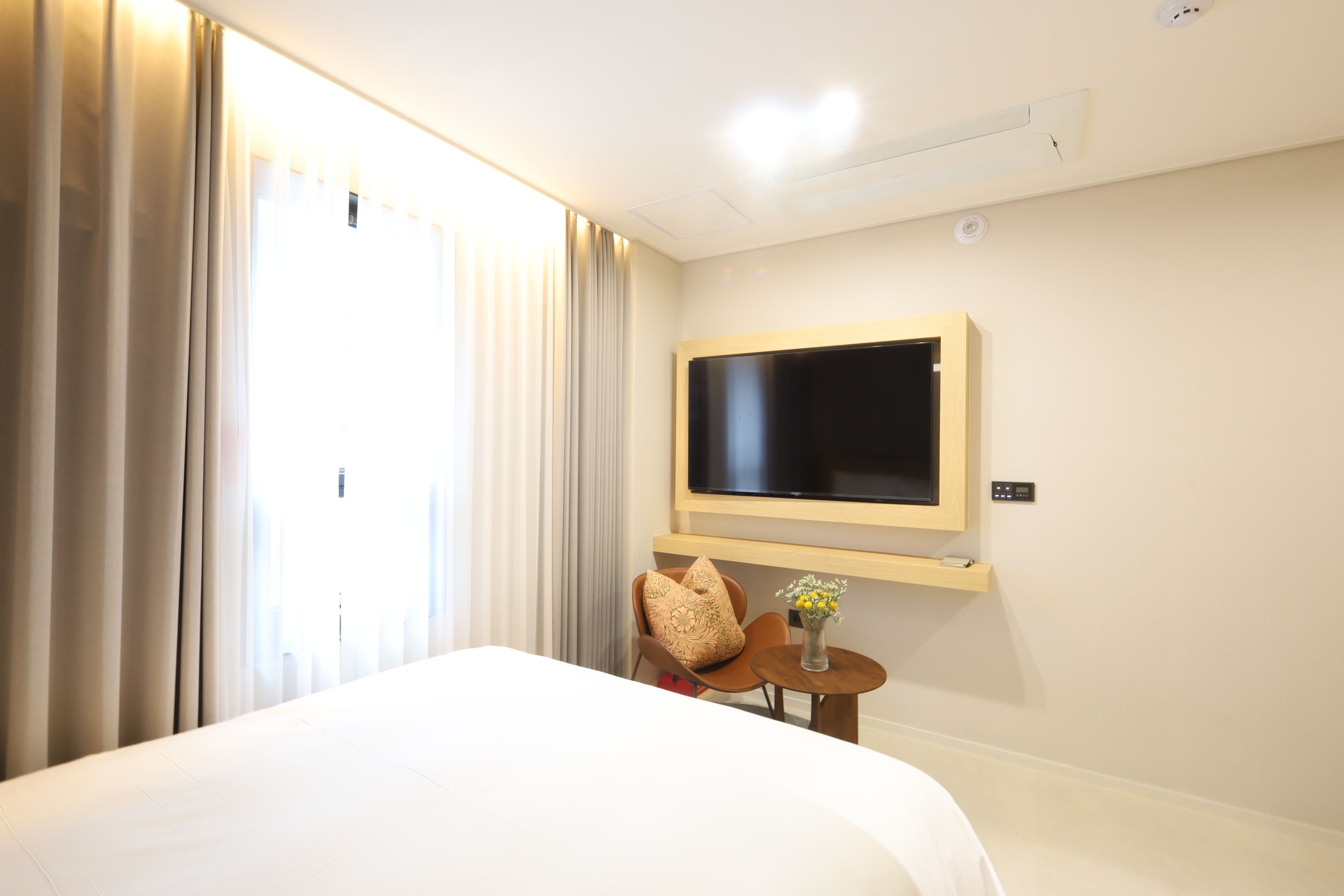 A hotel room with a bed, a TV mounted on the wall, a small table with a vase, and a chair with a decorative pillow.