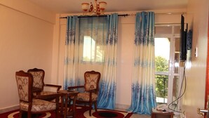 A room with a dining table and four chairs, a small side table with a telephone, and a window with blue curtains. - Latahzan guest house (Moyale)