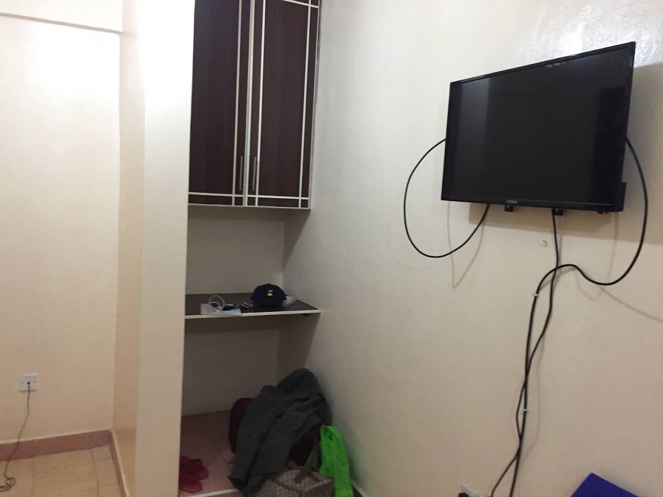 Ein kleines Zimmer mit an der Wand montiertem Fernseher, einem Fenster mit Gitterstäben und einem Regal mit diversen Gegenständen.