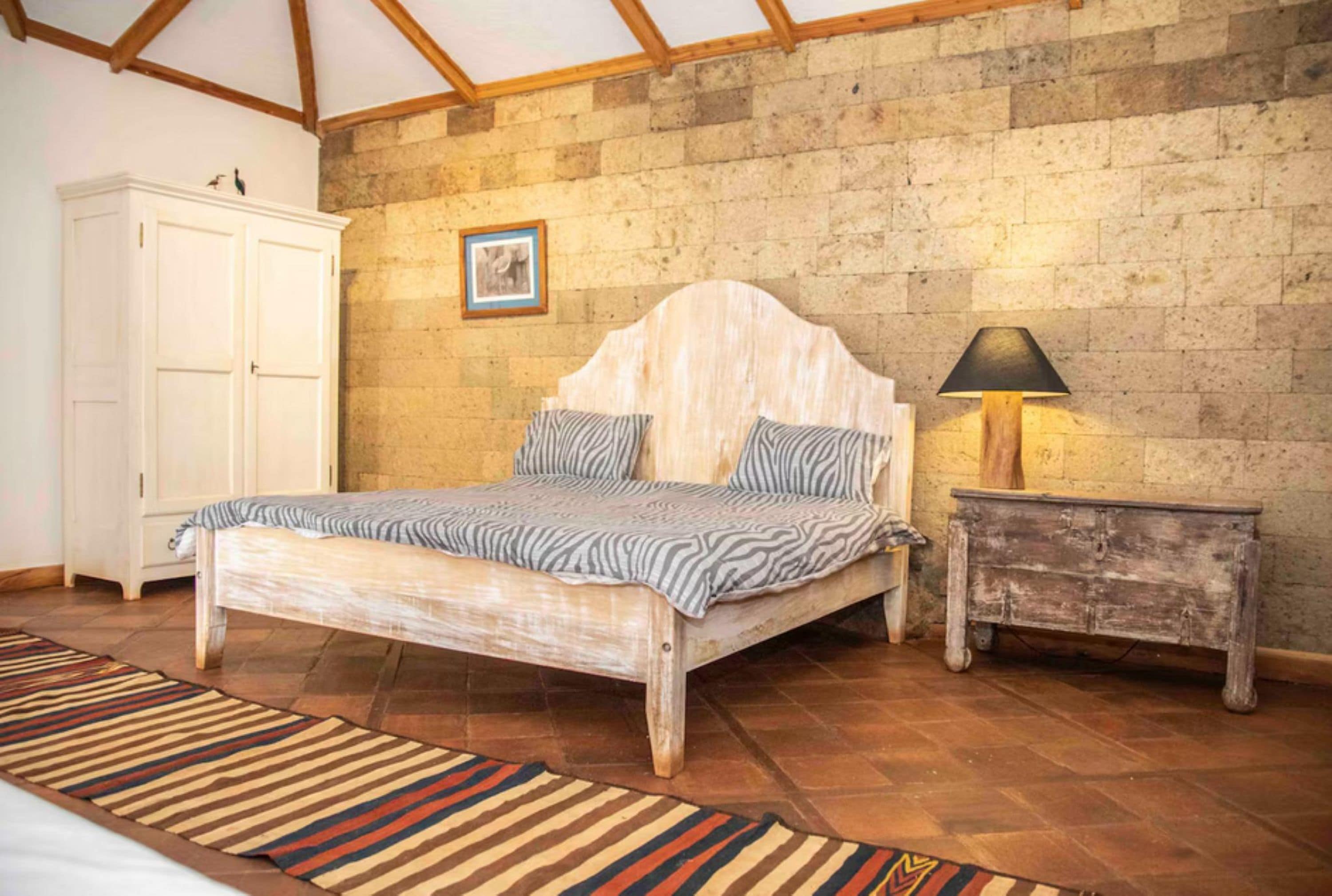 A bedroom with a wooden bed, a white wardrobe, a small bedside table with a lamp, and a stone wall.