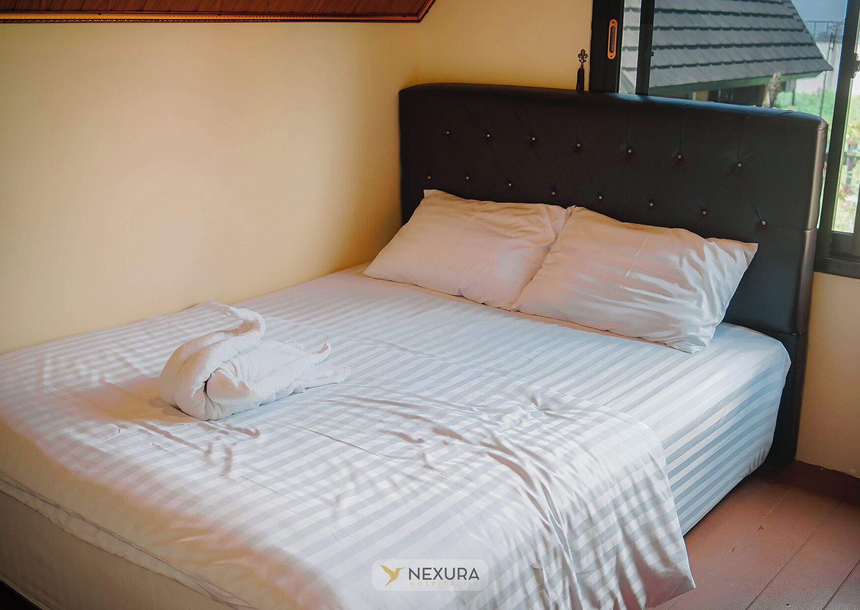 A neatly made bed with striped bedding and two pillows, a dark headboard, and a window with a view of a house.