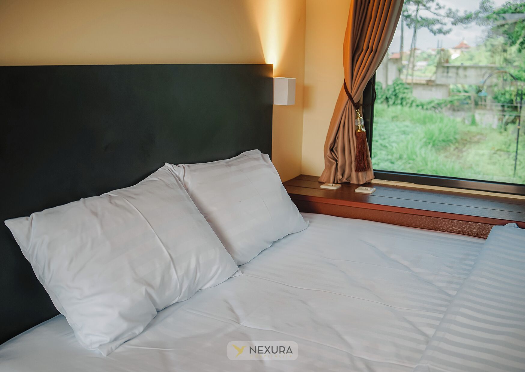 Una cama bien hecha con sábanas blancas en una habitación con cabecera oscura, una ventana con cortinas y vistas al exterior lleno de vegetación.