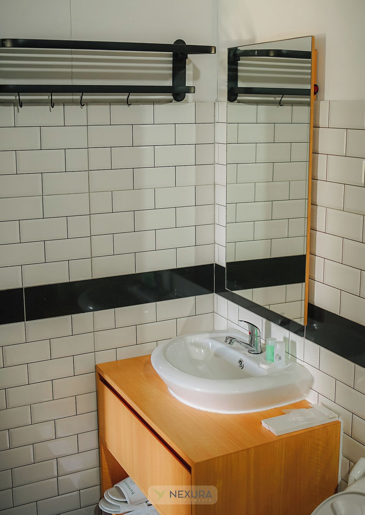 Une salle de bain avec des murs recouverts de carreaux blancs, un meuble-lavabo en bois, un lavabo blanc avec un miroir au-dessus, et un porte-serviettes.