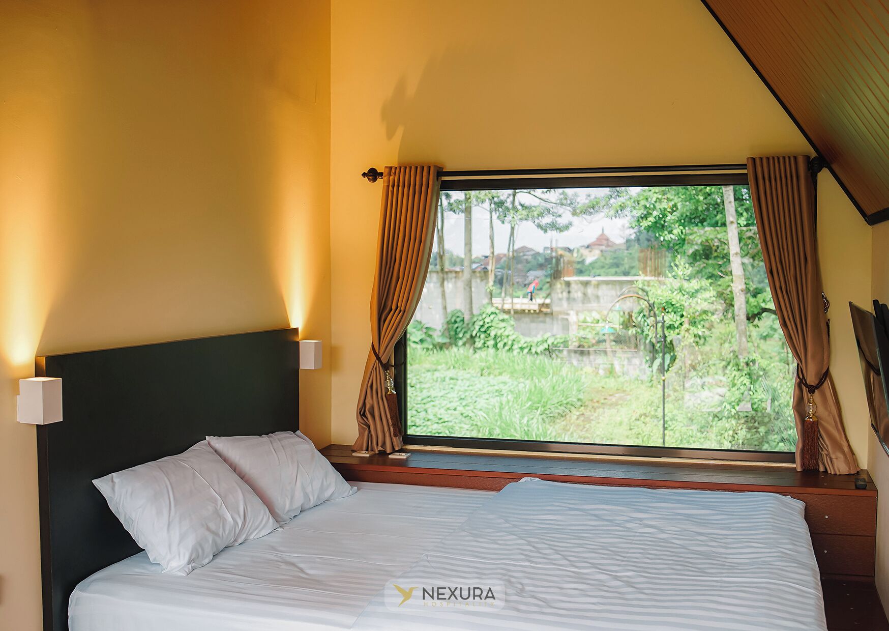 Une chambre d’hôtel avec un lit, une fenêtre avec des rideaux, une table de chevet et une lampe fixée au mur.