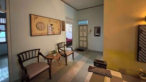Un couloir avec un sol à carreaux, deux chaises en bois, une petite table avec une plante et un mur orné de tableaux et d’une horloge.