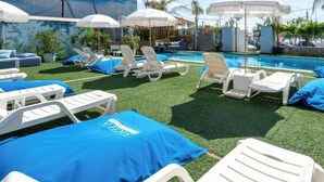 Zona junto a la piscina con sillas de descanso blancas y toallas azules, rodeada de sombrillas y palmeras.