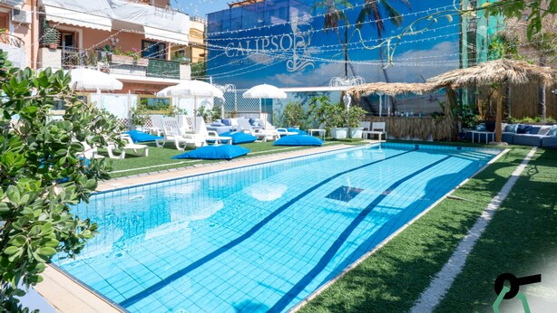 Una piscina con sillas de descanso y sombrillas, rodeada de follaje y un edificio con un mural azul.