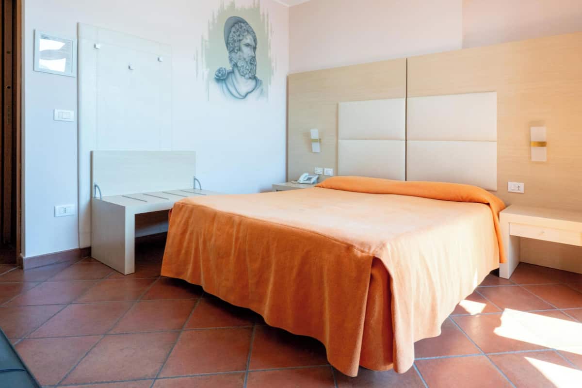 A hotel room with a bed, a desk, a chair, a wall-mounted phone, and a painting.