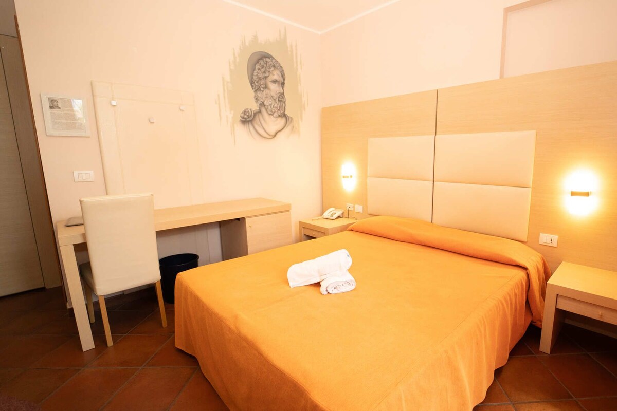 Een hotelkamer met een bed, bureau, stoel en een schilderij aan de muur.