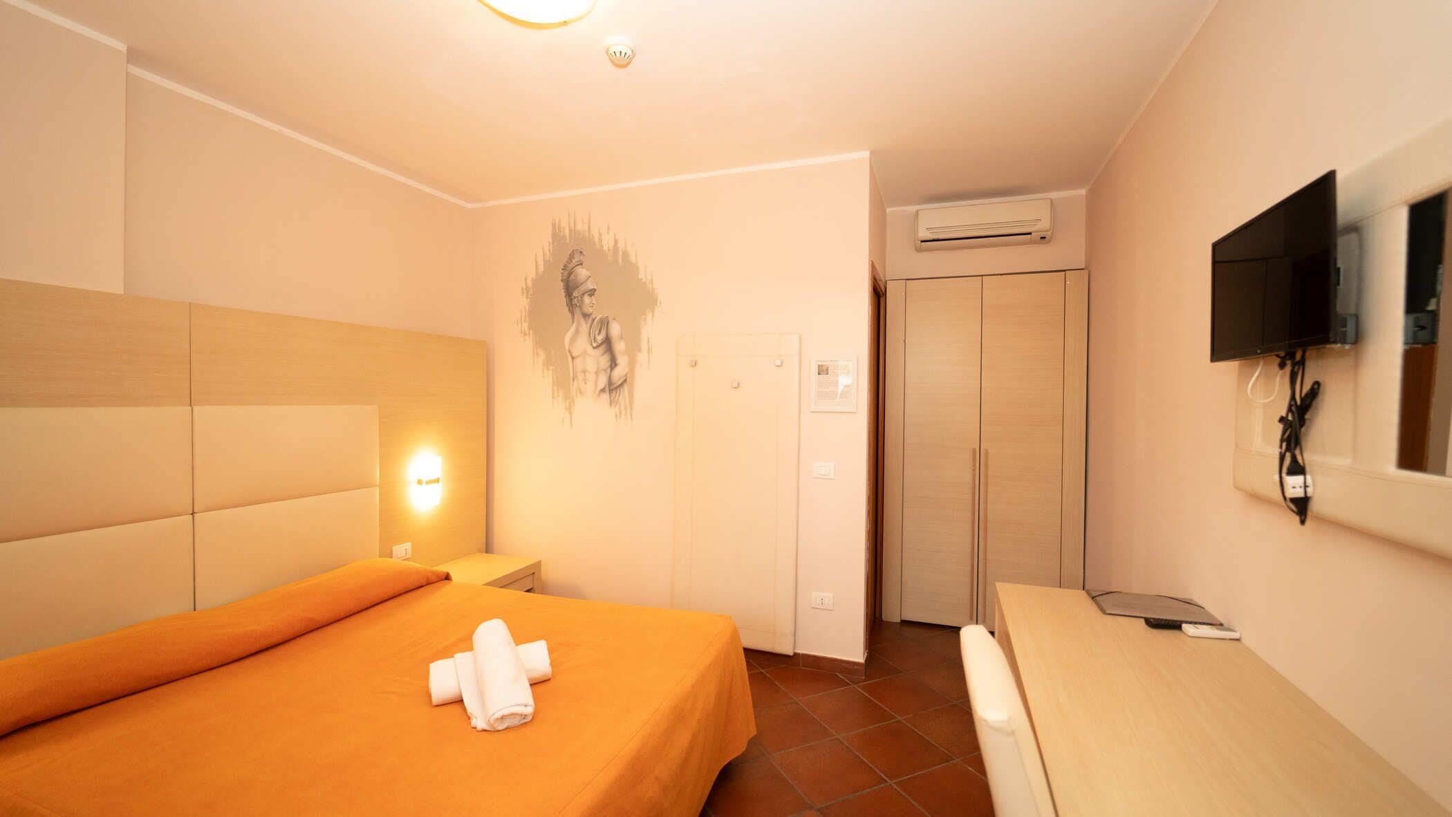 Een hotelkamer met een bed, nachtkastje, een aan de muur bevestigde televisie, airconditioning en een deur.