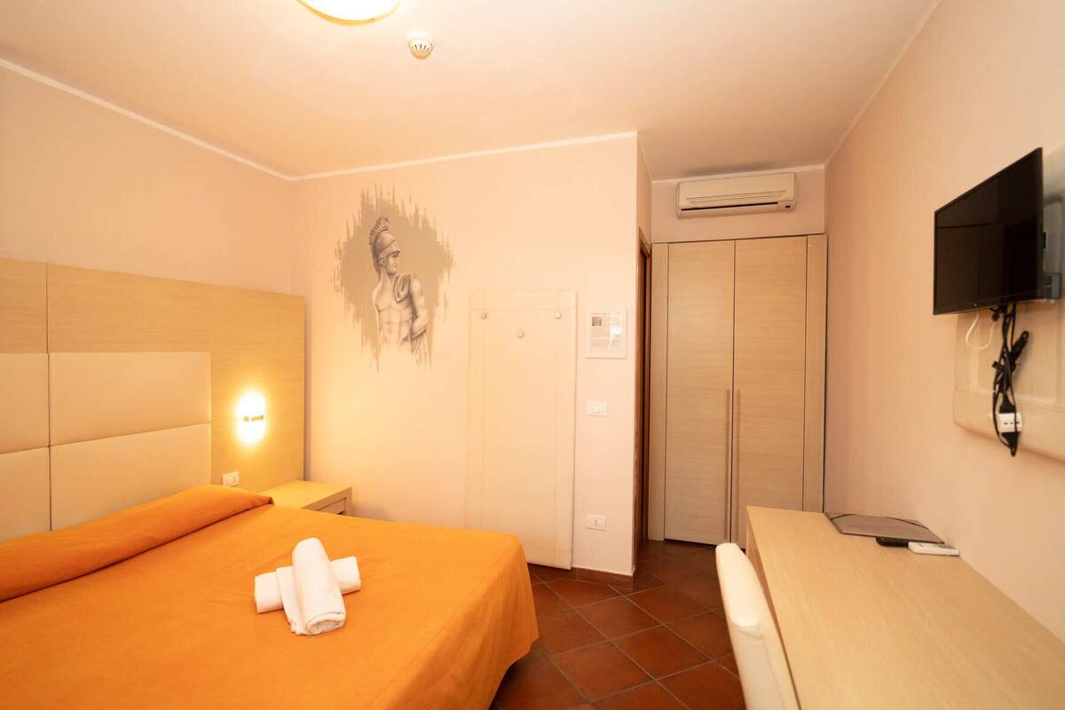 A hotel room with a bed, a bedside table, a wall-mounted TV, an air conditioning unit, and a door.