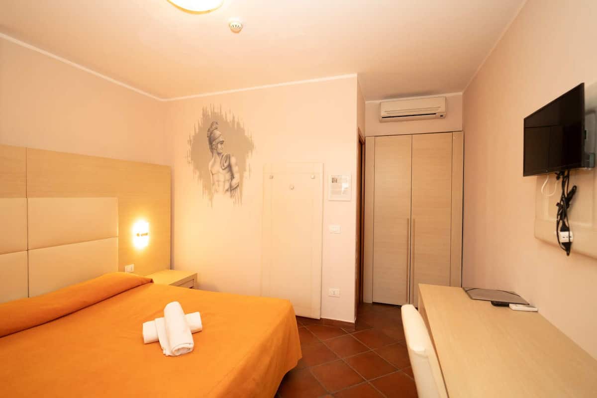 A hotel room with a bed, a bedside table, a wall-mounted TV, an air conditioning unit, and a door.
