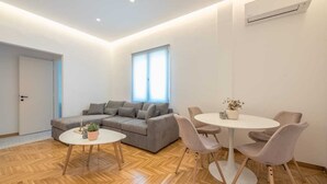 Ein modernes Wohnzimmer mit einem grauen Sofa, einem runden Tisch mit Stühlen, einem Blumentopf und einem Fenster mit Jalousien.