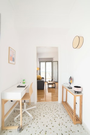 Ein modernes, minimalistisches Zimmer mit einem weißen Schreibtisch, einem Stuhl, einem kleinen Tisch mit Lampe und einem gerahmten Bild an der Wand.