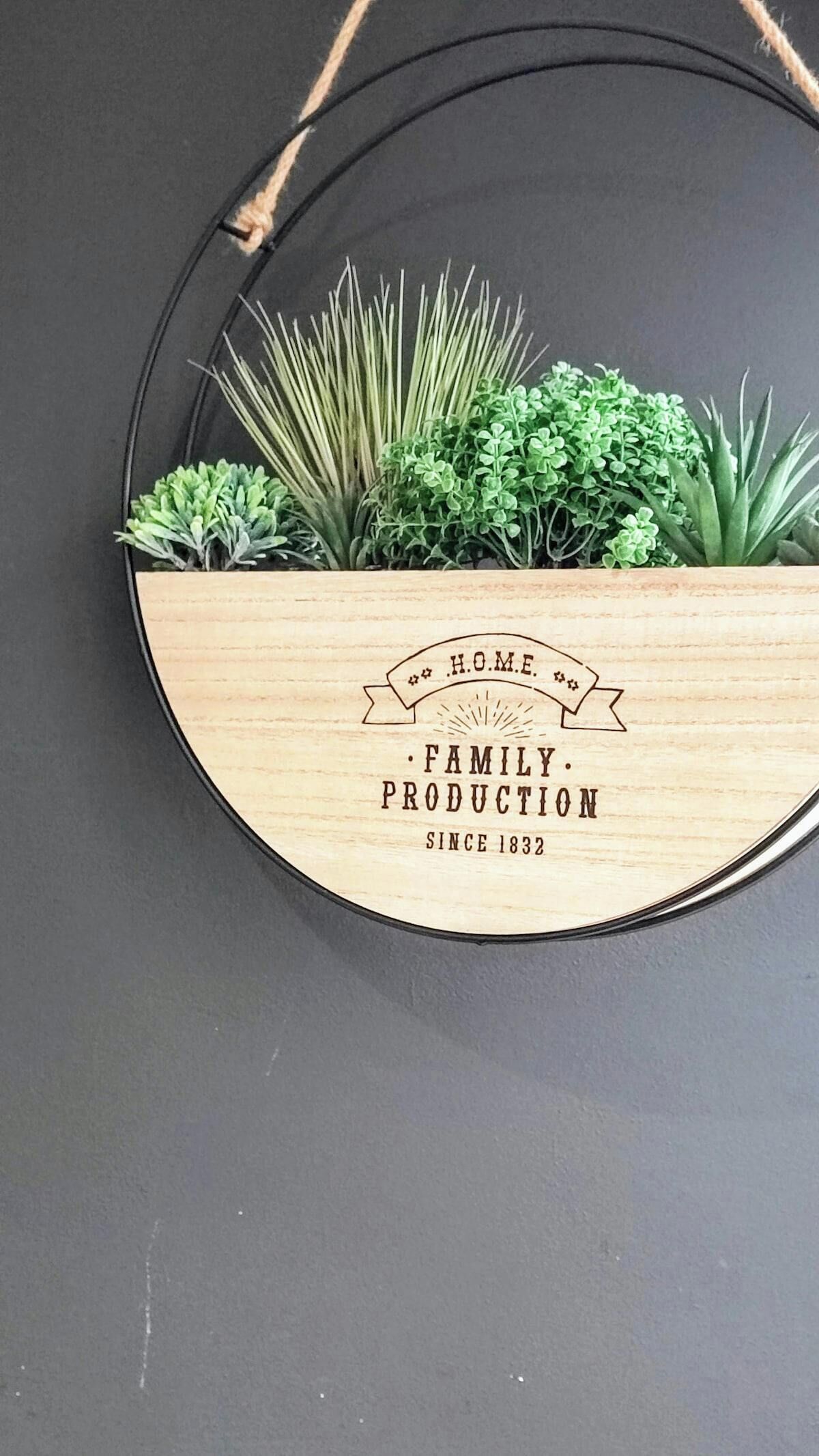 Um vaso de madeira circular com uma alça preta, contendo diversas plantas verdes. Possui uma etiqueta com a inscrição “HOME FAMILY PRODUCTION SINCE 1832”.