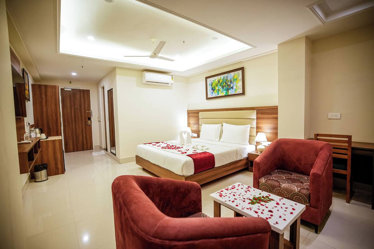 A hotel room with a bed, two red armchairs, a small table with rose petals, a ceiling fan, and an air conditioning unit.