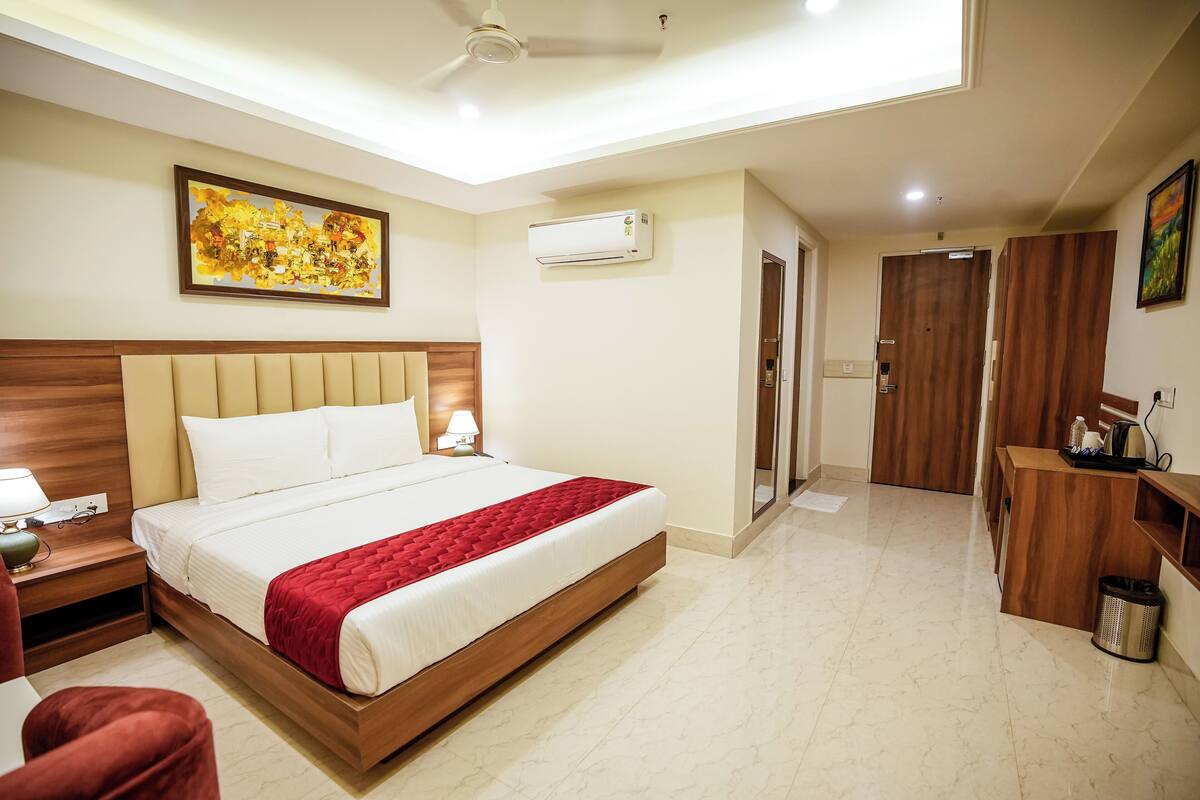 A hotel room with a bed, bedside table, lamp, air conditioning unit, wardrobe, and a painting on the wall.