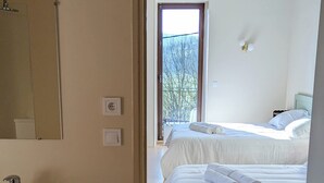 A bedroom with two beds, a bathroom with a sink, and a large window providing a view of trees. - Domaine la Pause in Aveyron, country house - ideal for families or friends (Vabres-l'Abbaye)