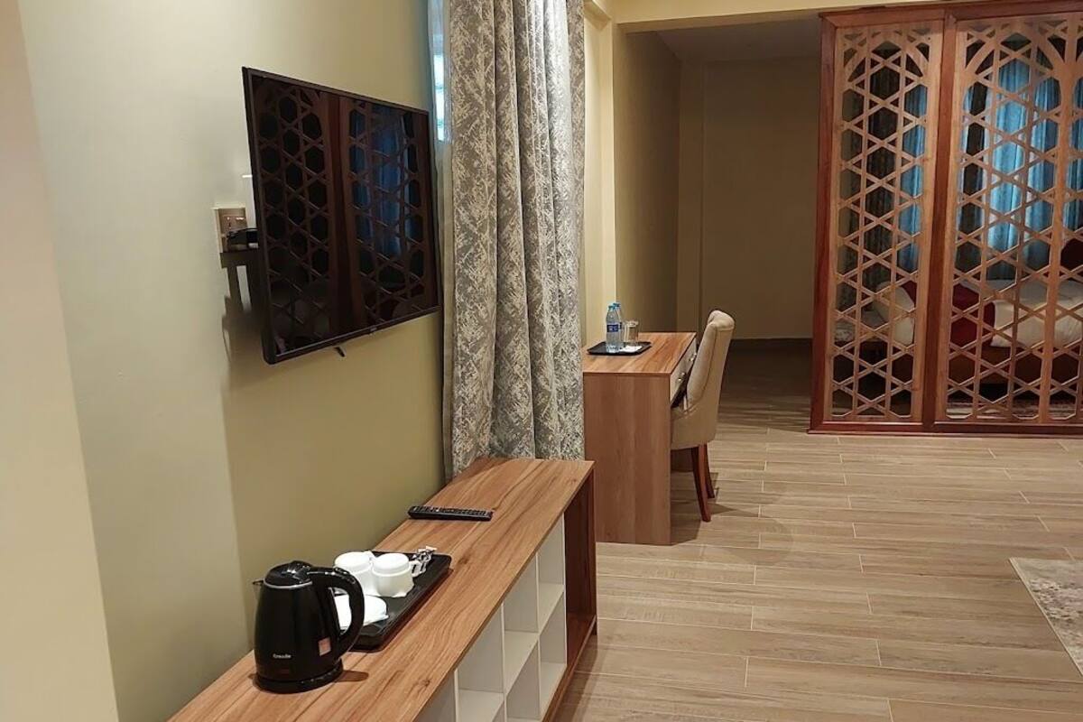 A hotel room with a wooden console table, a television mounted on the wall, a desk with a chair, and a decorative wooden partition.