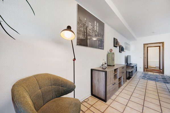 Una sala de estar con sofá, una lámpara de pie, un mueble con televisor y un cuadro de un paisaje urbano en la pared.