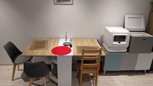 A dining area with a wooden table, two chairs, a red placemat, a framed picture on the wall, and a kitchenette with a refrigerator and microwave. - Chez steph (Le Puy-en-Velay)