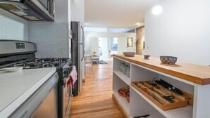 A modern kitchen with a gas stove, microwave, and oven. There is a wooden shelf with kitchen utensils and a bowl. The kitchen has a wooden floor and white walls. - XL Prime Tribeca Furnished Loft (New York)