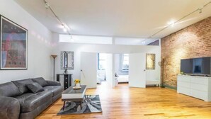 A modern living room with a leather sofa, a coffee table, and a flat-screen TV against a brick wall. - XL Prime Tribeca Furnished Loft (New York)