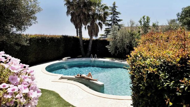 Une piscine où une personne se détend au bord, entourée de différentes plantes et arbres.