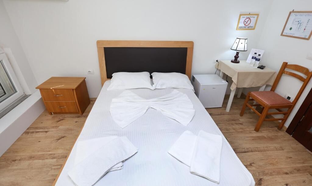 Una cama bien hecha con sábanas blancas, una mesita de noche de madera, un escritorio con una lámpara y una silla.