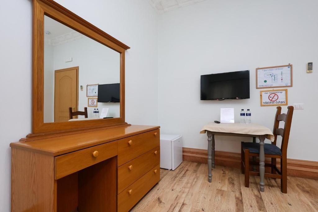 Quarto com cômoda de madeira, escrivaninha com cadeira, TV de tela plana na parede e um eletrodoméstico pequeno e branco.