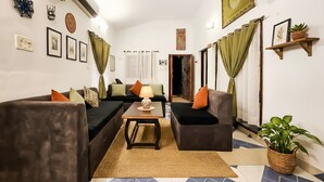 A living room with two sofas, a coffee table, and a lamp. There are wall decorations, a plant, and a shelf with a plant. - The Plix Villa (Vagator)