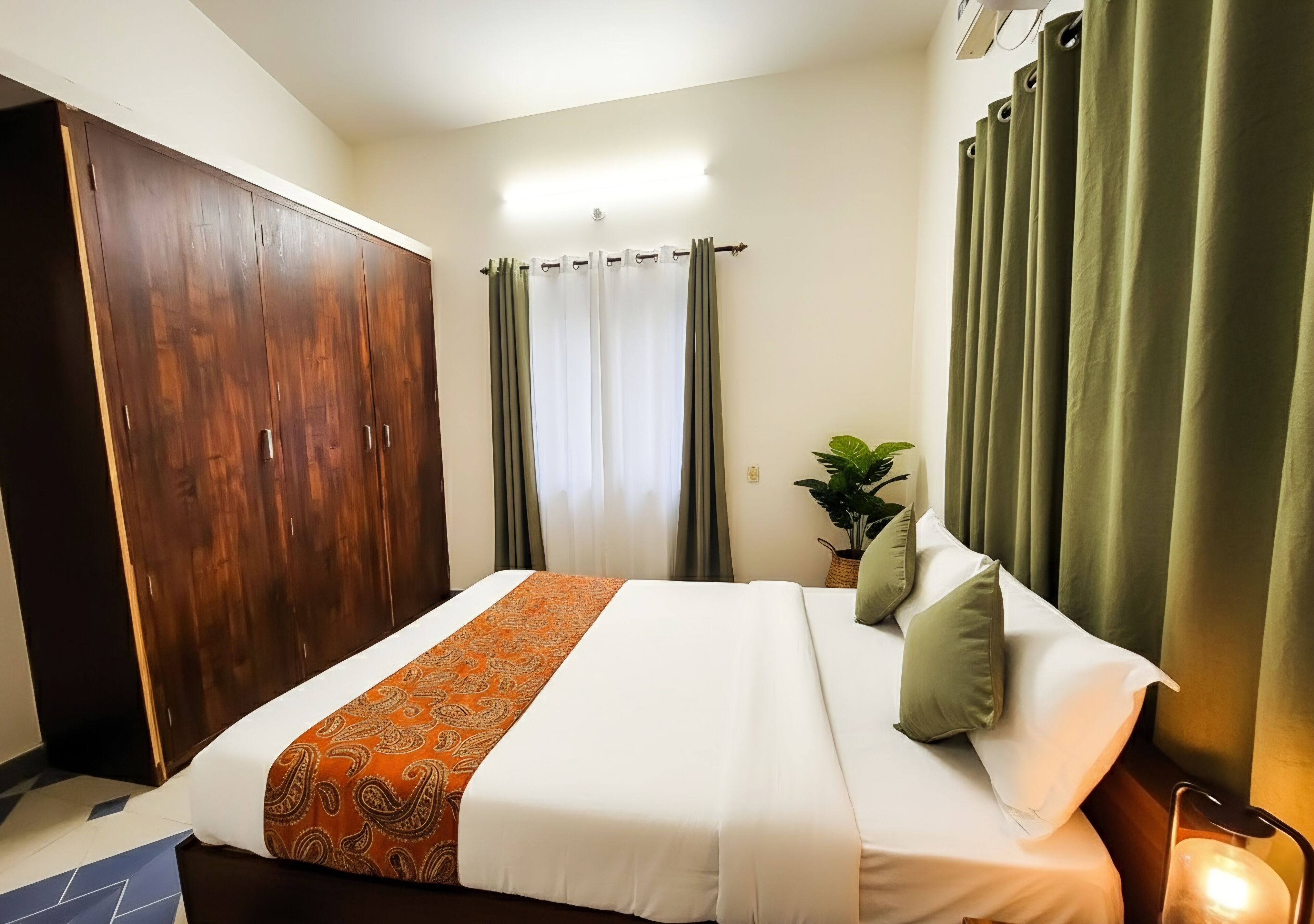 Une chambre d’hôtel avec une grande armoire, un lit avec une literie blanche et un couvre-lit à motifs orange, des rideaux verts et une plante sur la table de chevet.