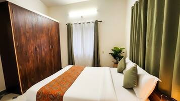 Une chambre d’hôtel avec une grande armoire, un lit avec une literie blanche et un couvre-lit à motifs orange, des rideaux verts et une plante sur la table de chevet.