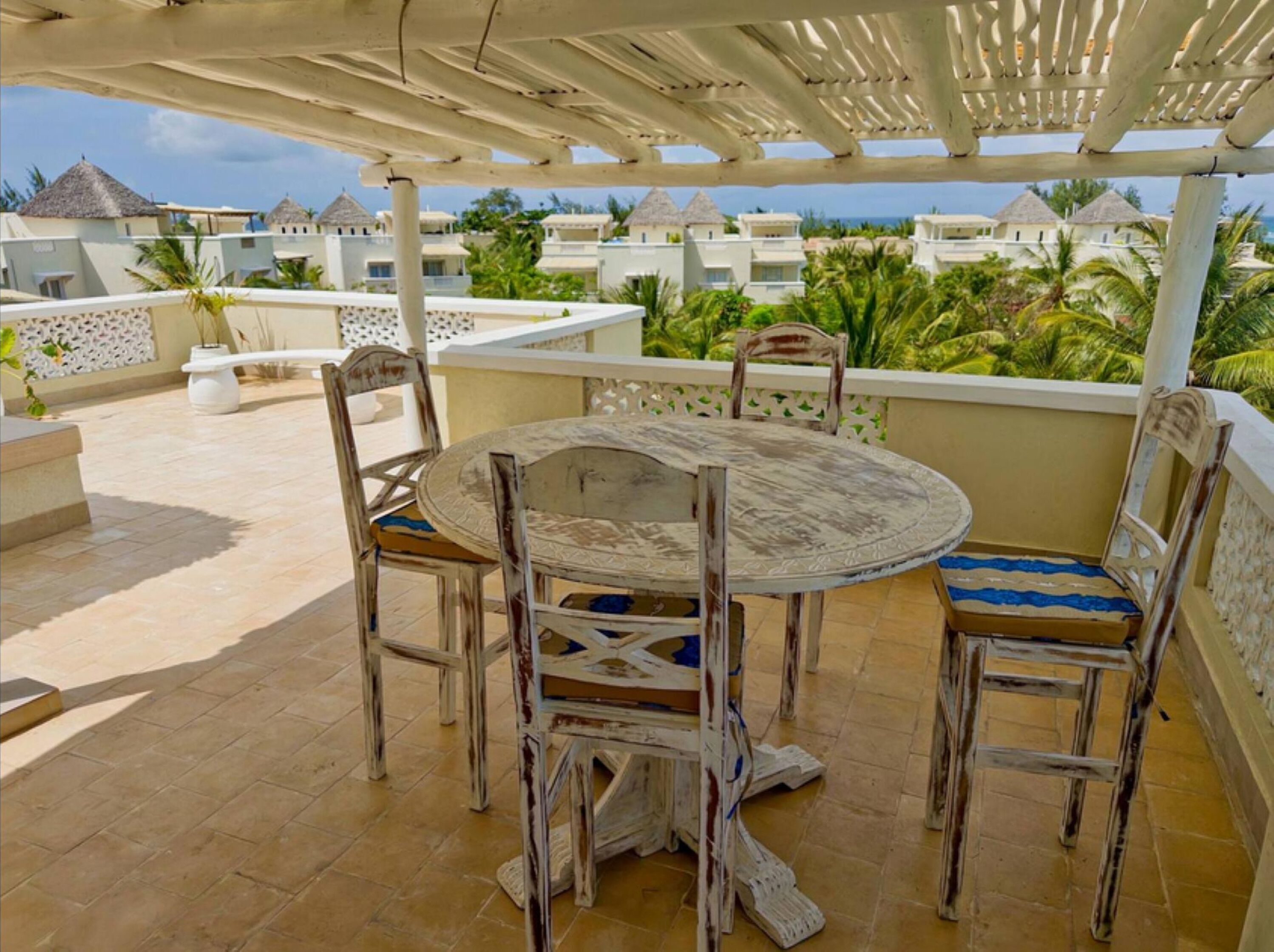Une table ronde entourée de chaises, placée sous une pergola, d’où l’on peut admirer un complexe hôtelier avec des palmiers et des bâtiments.