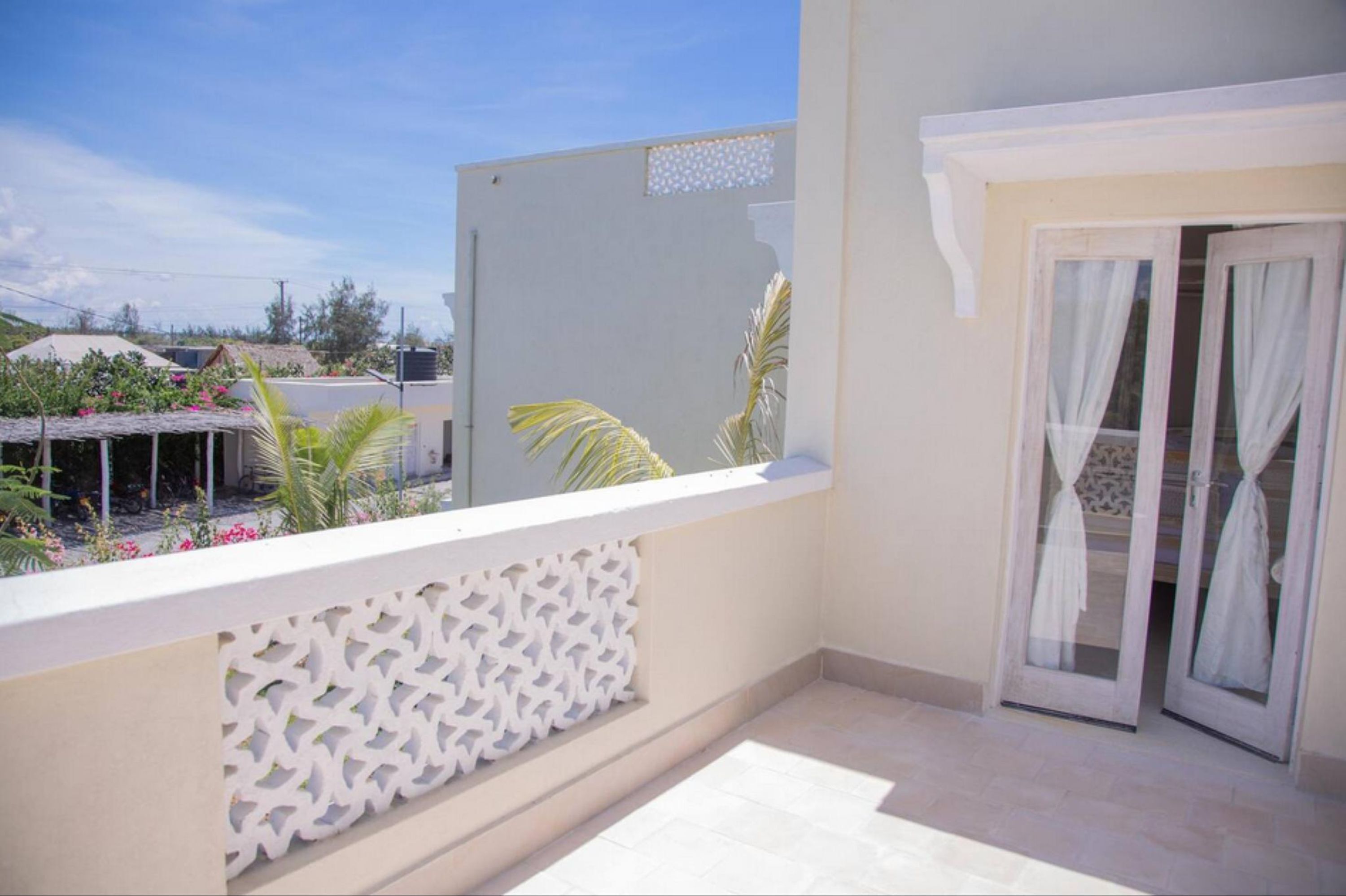 A balcony with a white decorative panel, a sliding glass door with white curtains, and a view of neighboring buildings and greenery.
