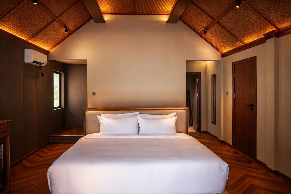 A hotel room with a large bed, wooden flooring, a sloped ceiling with recessed lighting, a window, and a door.