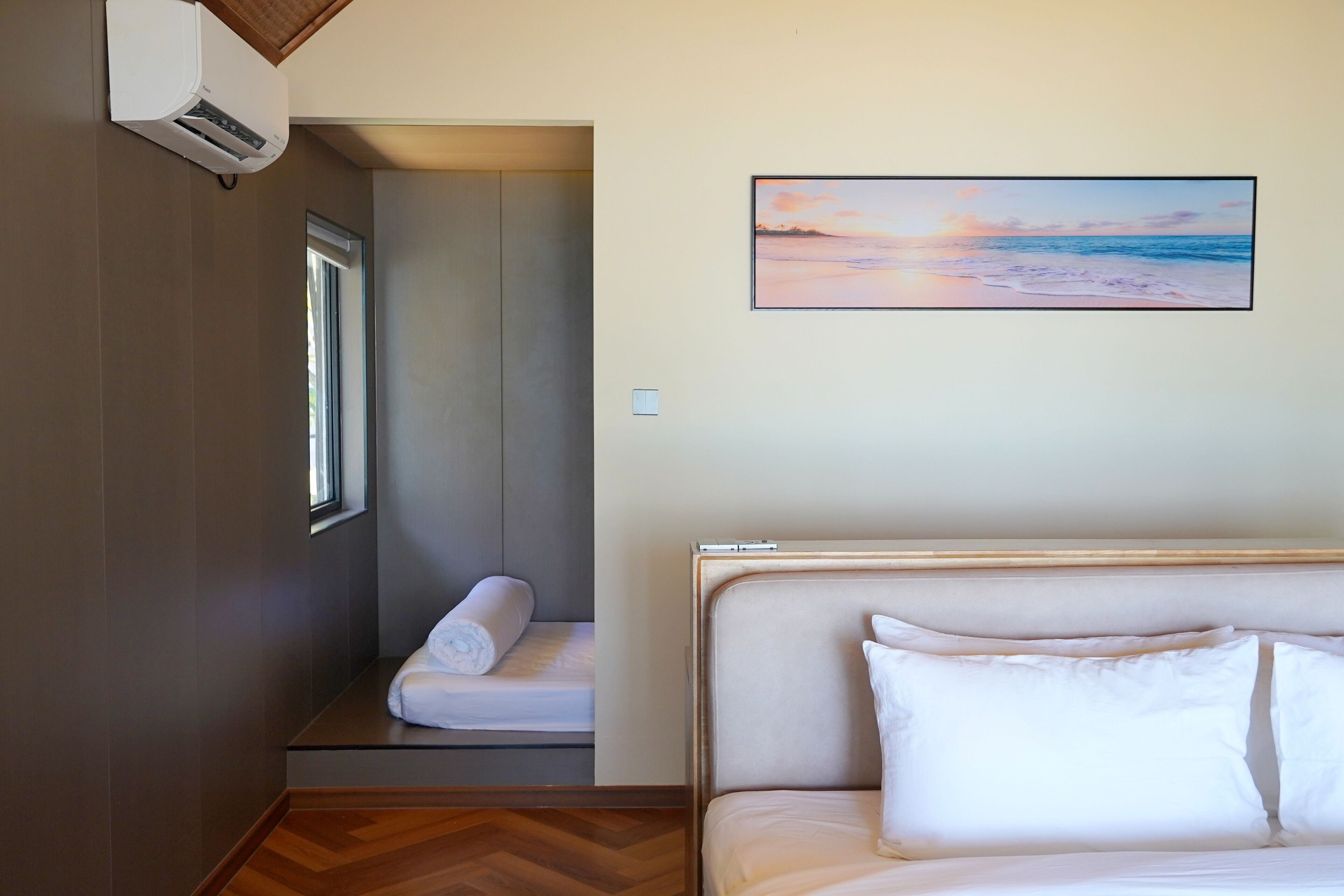 A bedroom with a bed, a window, a wall-mounted air conditioner, and a painting of a beach scene.